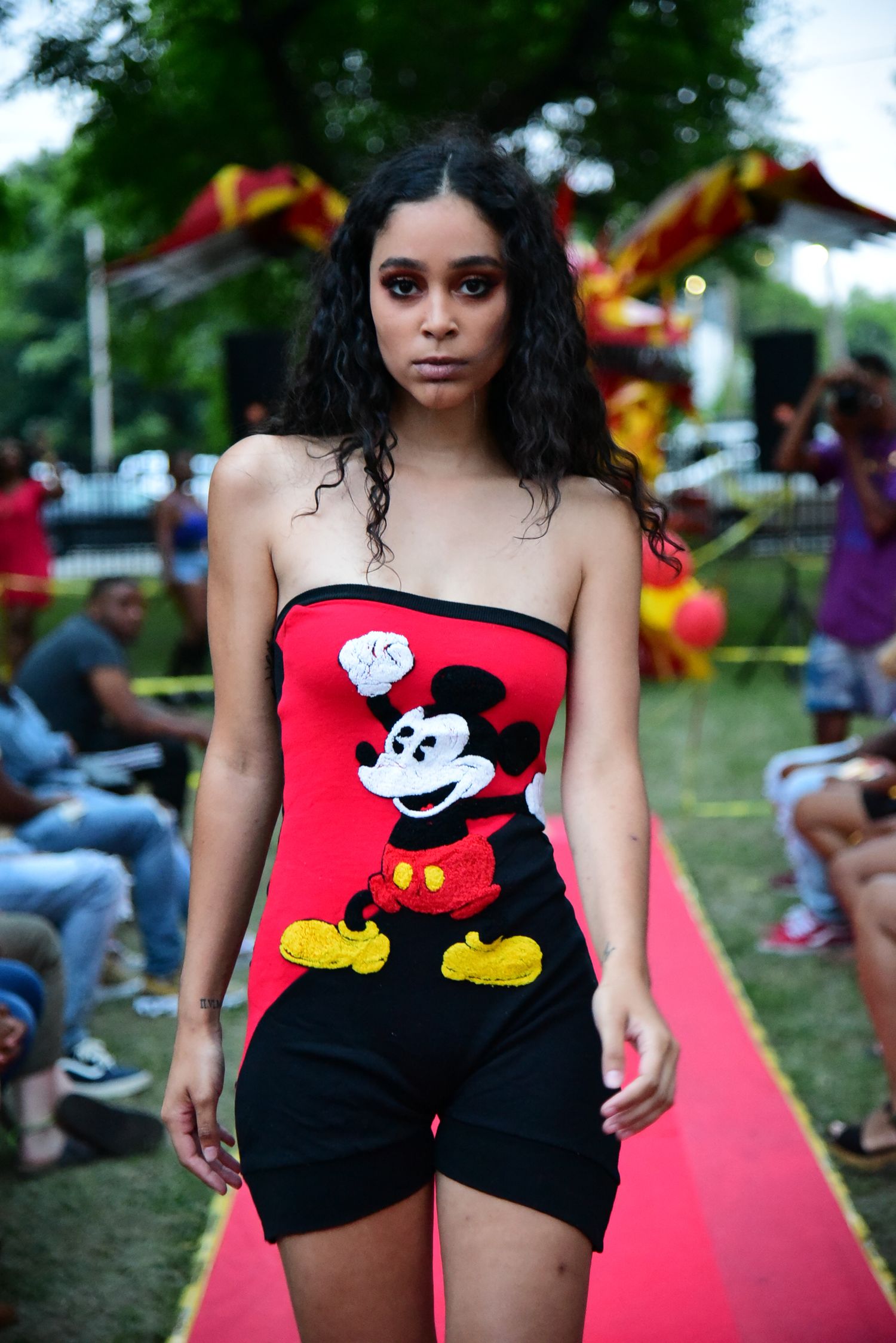 Model walks runway in Mickey Mouse themed outfit at outdoor fashion show.