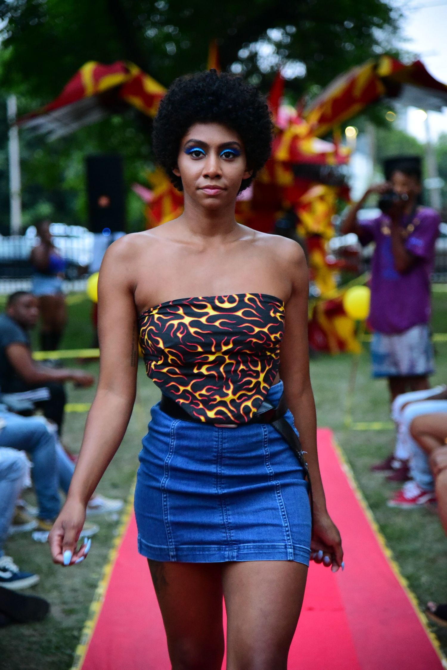 Model walks red carpet in patterned strapless top and denim skirt at outdoor fashion show.