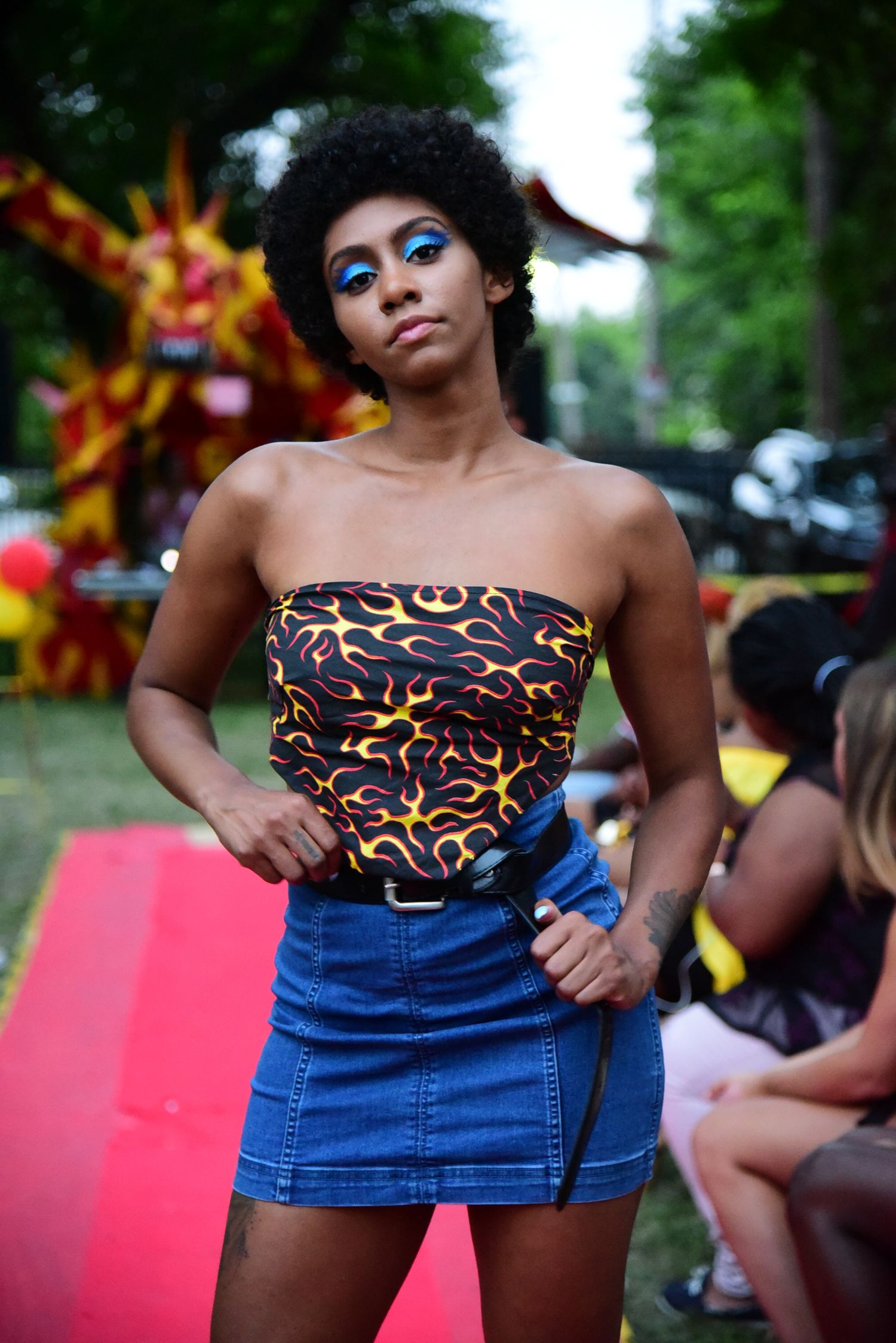Model in patterned top and denim skirt poses on red carpet at outdoor event.