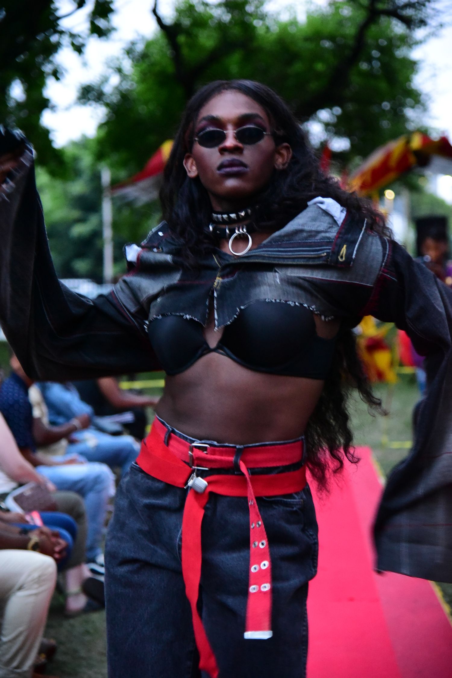 Fashion model wearing black crop top with red belt poses on outdoor runway with crowd in background.