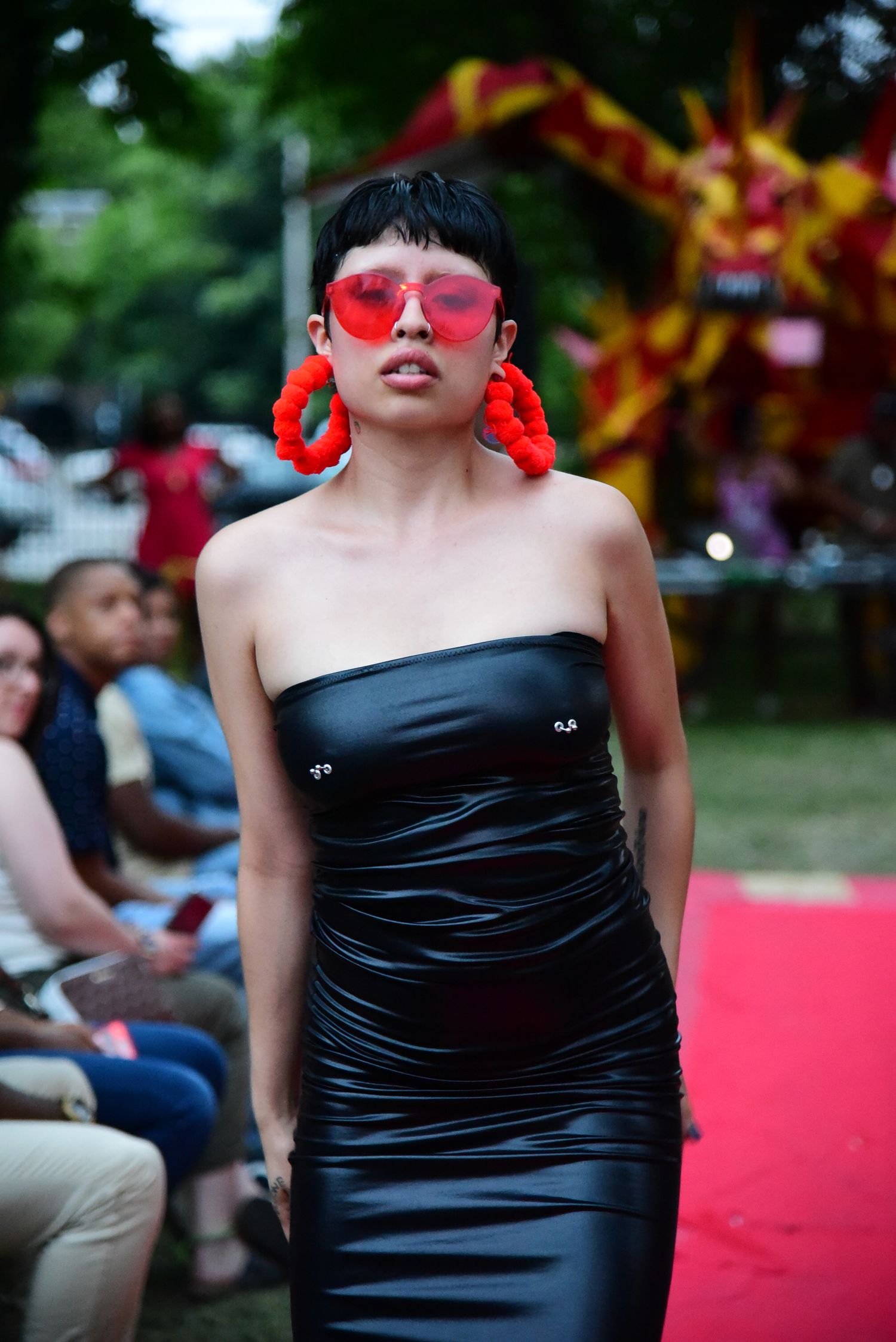 Model in black strapless dress and red sunglasses walks runway at outdoor fashion event.