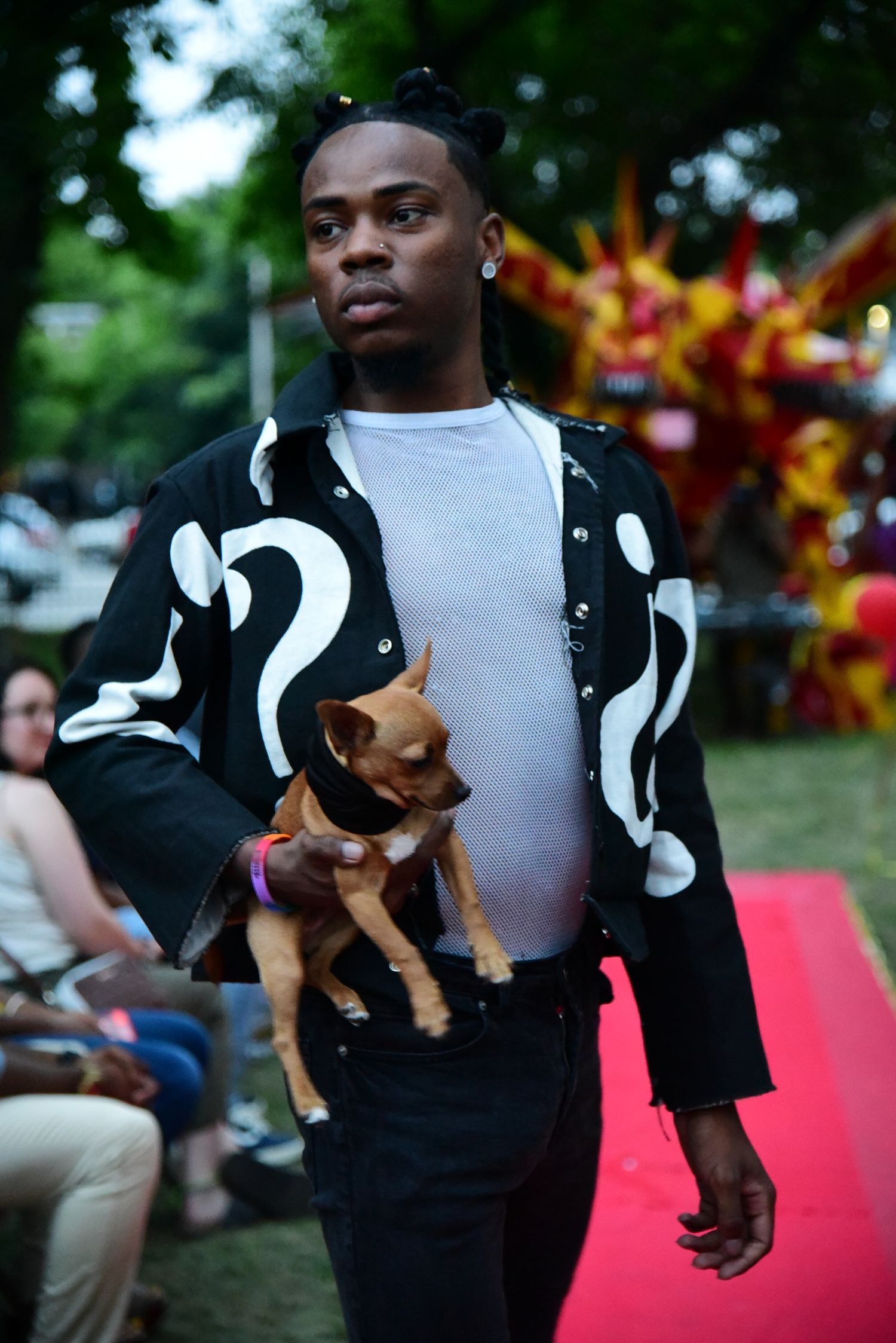 Model in black and white jacket carries small dog while walking red carpet at outdoor event.