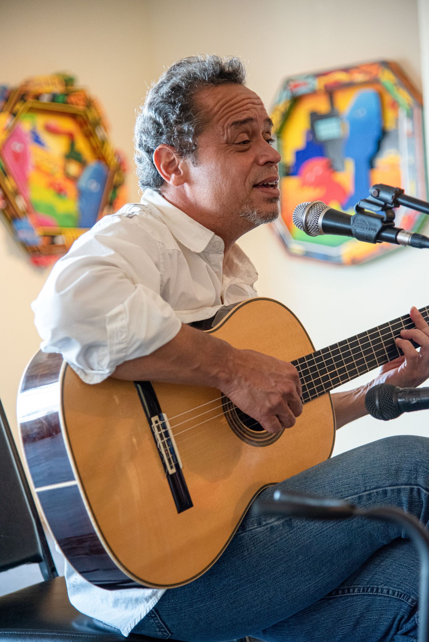 Classical guitarist performing with acoustic guitar against wall decorated with colorful art pieces.