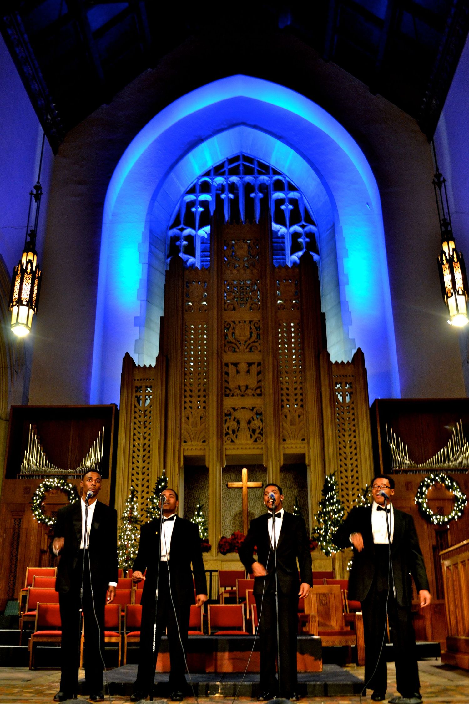 Four performers in tuxedos singing on stage in church with illuminated blue Gothic arch and decorative altar.