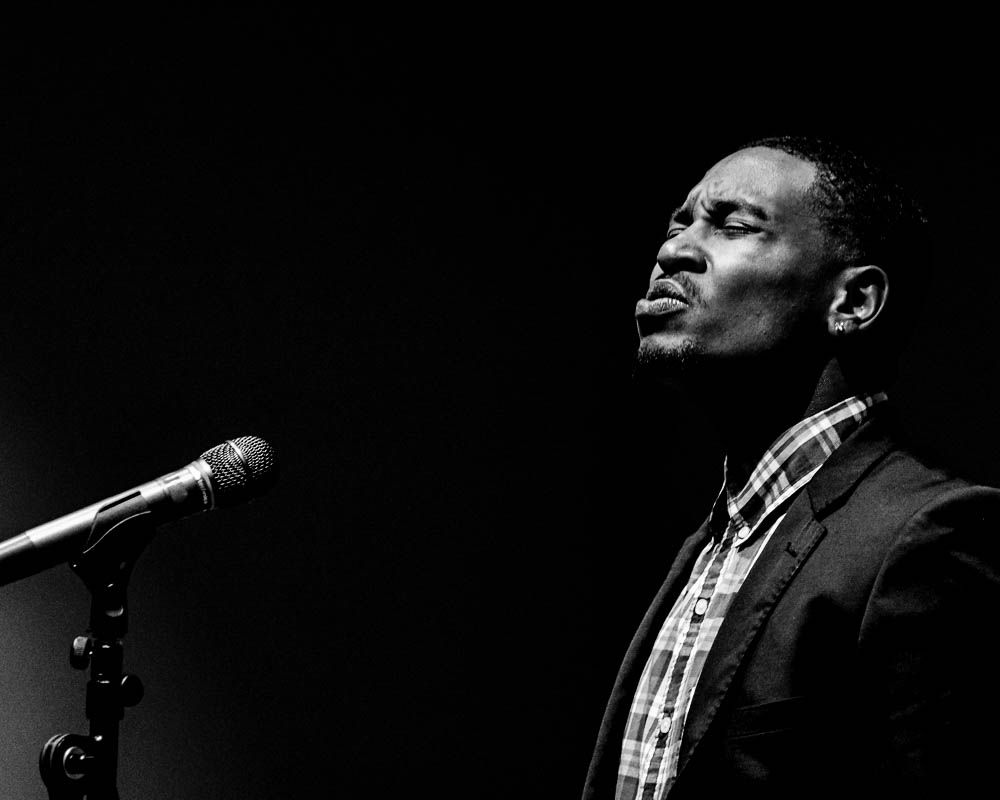 Dramatic black and white side profile of performer singing into microphone on dark stage.