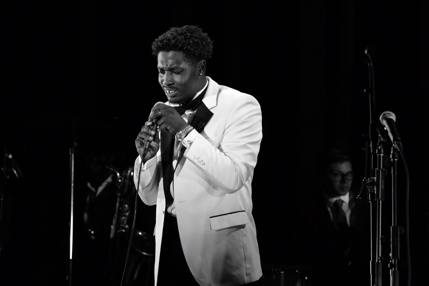 Performer in white tuxedo jacket singing into microphone against dark background in classic black and white style.