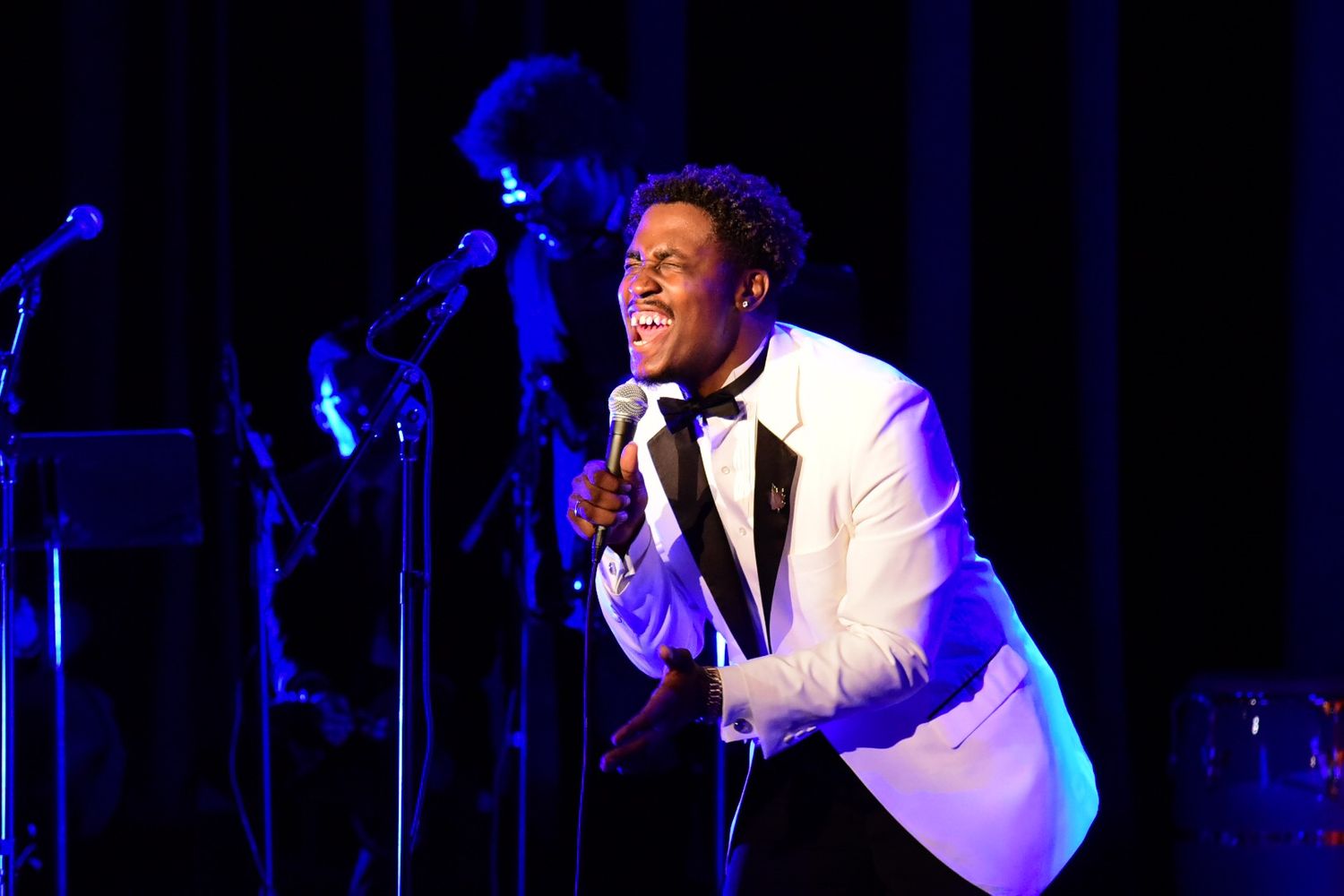 Performer in white tuxedo energetically singing on stage with blue lighting and band in background.