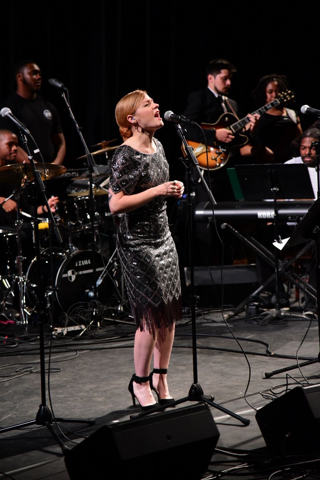 Singer in sequined dress performs with full band on concert stage with instruments and equipment.