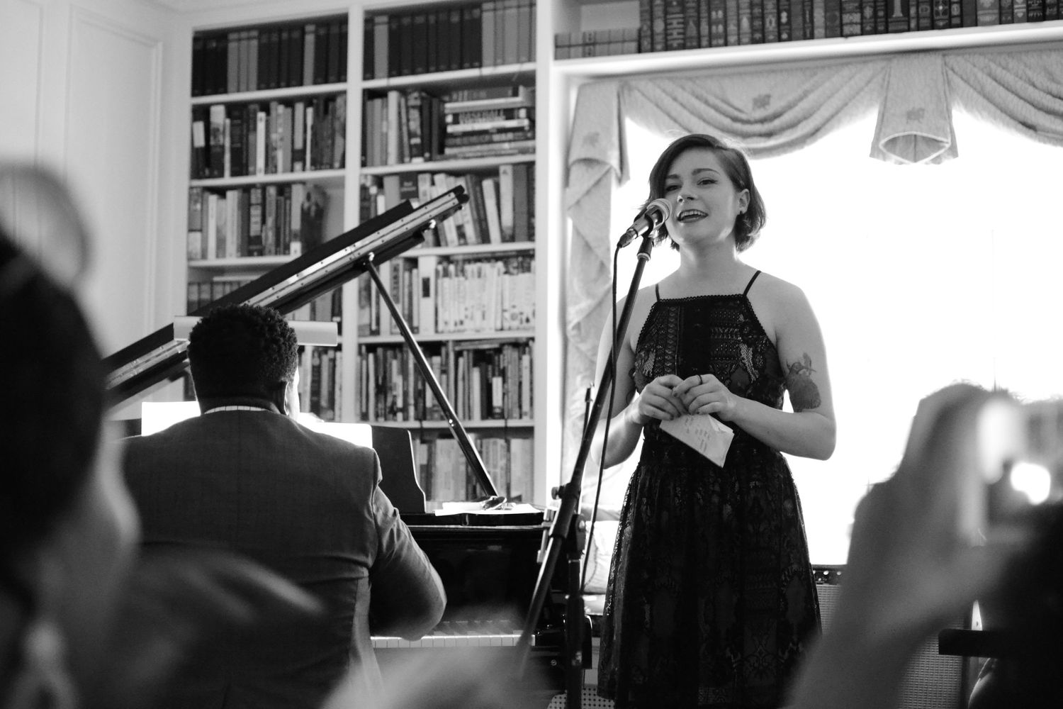 Black and white photo of performer singing in intimate library venue with microphone setup.