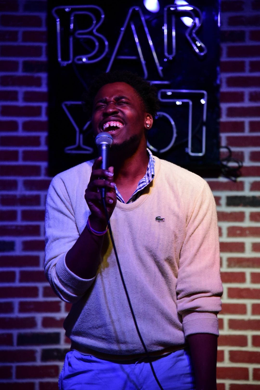 Performer singing at bar venue with neon sign and brick wall background in purple lighting.