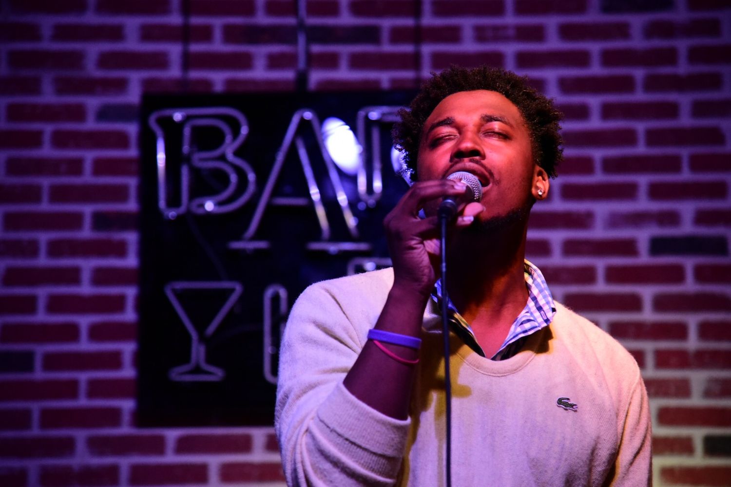 Singer performing passionately at bar venue with neon signage and brick wall backdrop.