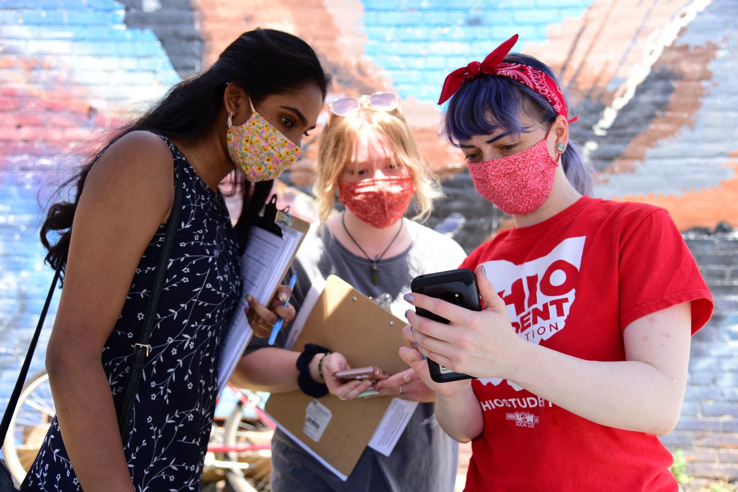 Group gathering outdoors wearing masks while discussing near brick wall.