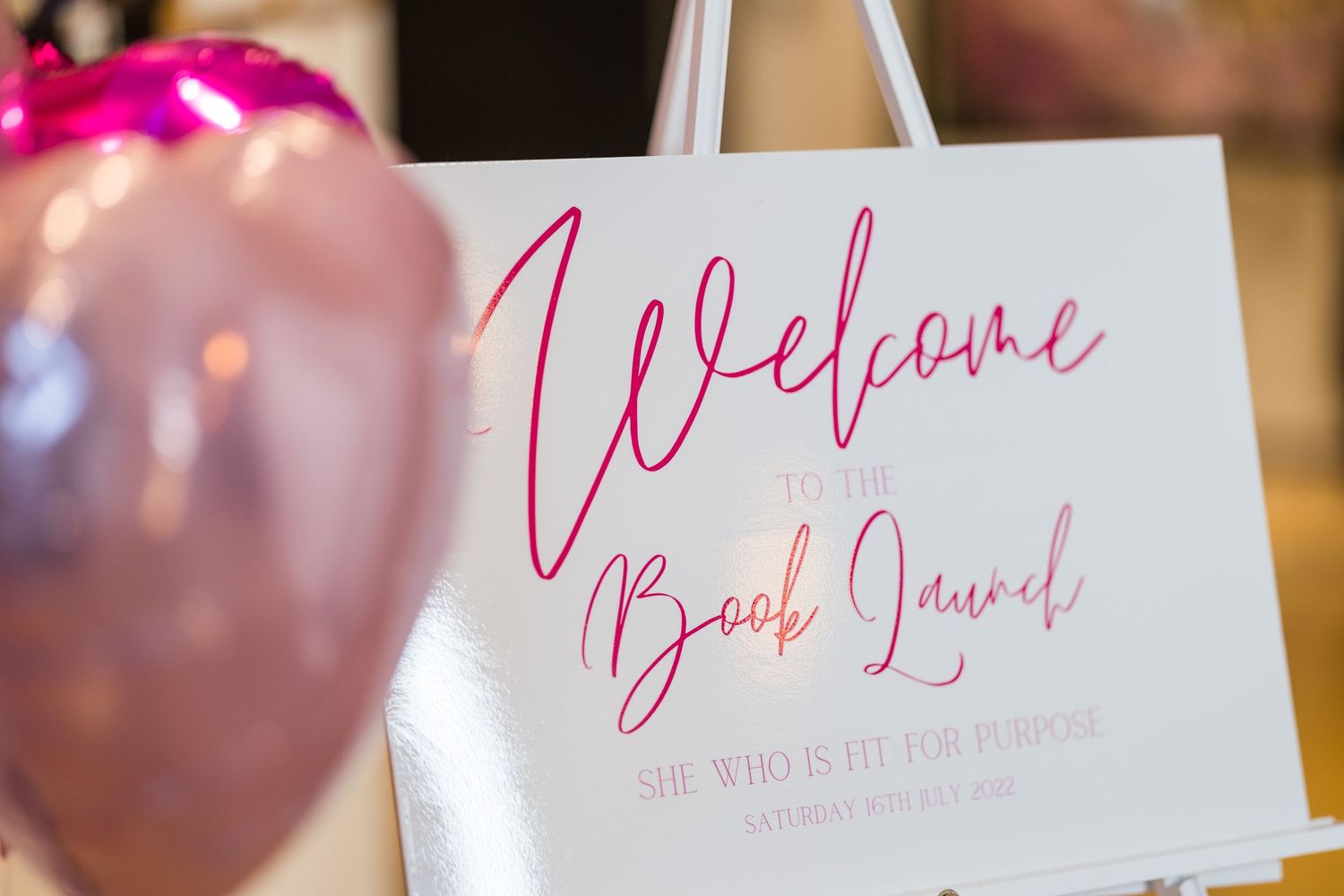 A welcome sign for a brand launch event with pink decor and balloons in the background.