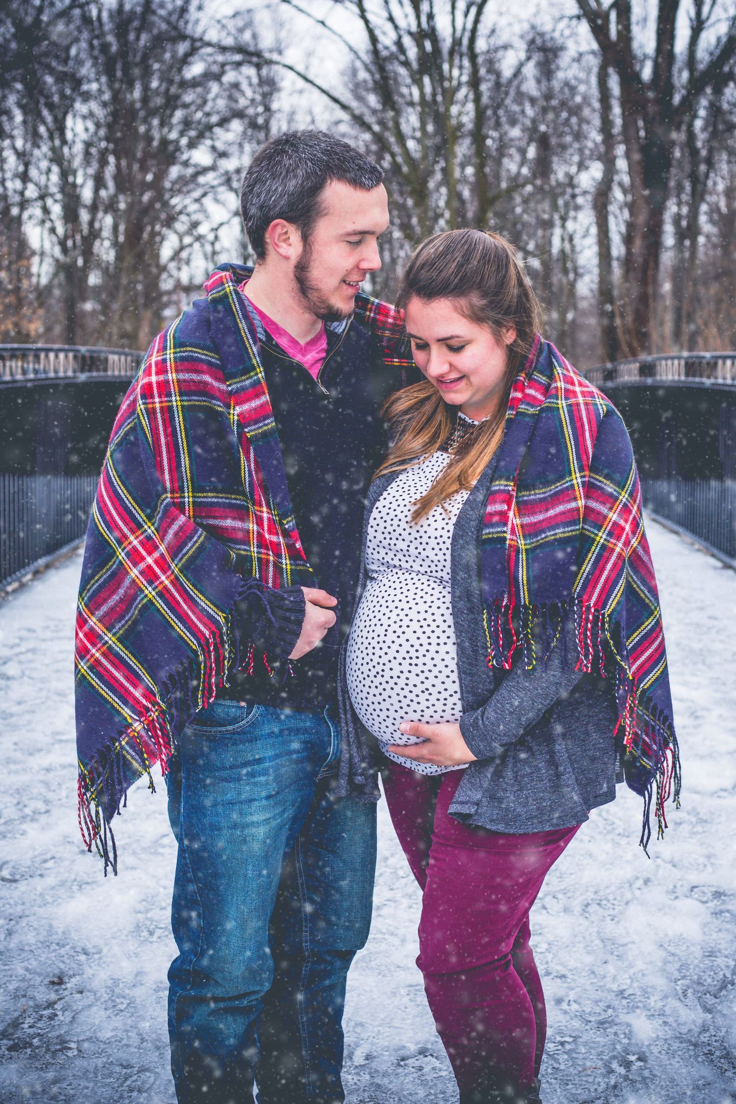 A couple wrapped in matching plaid blankets stands together in the snow.