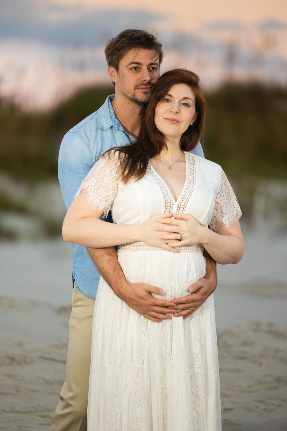 A couple embraces on a beach at sunset wearing a white lace dress and blue shirt.