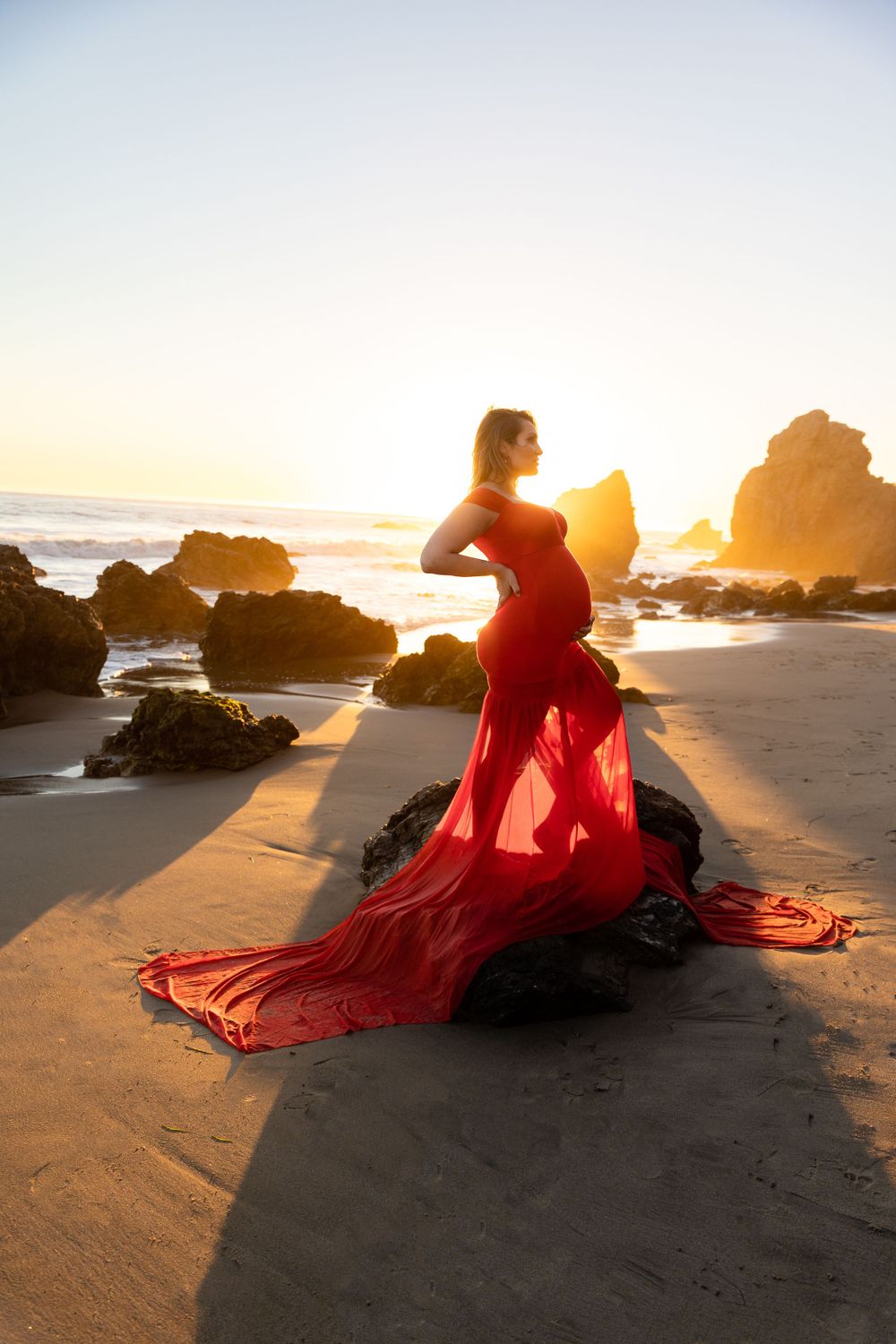 Silhouette of flowing red dress against golden sunset on beach with rocky outcrops.
