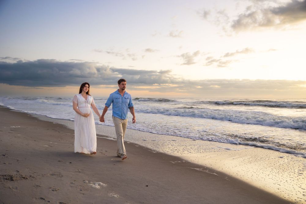 A couple walks along a sandy beach with waves crashing during sunset.