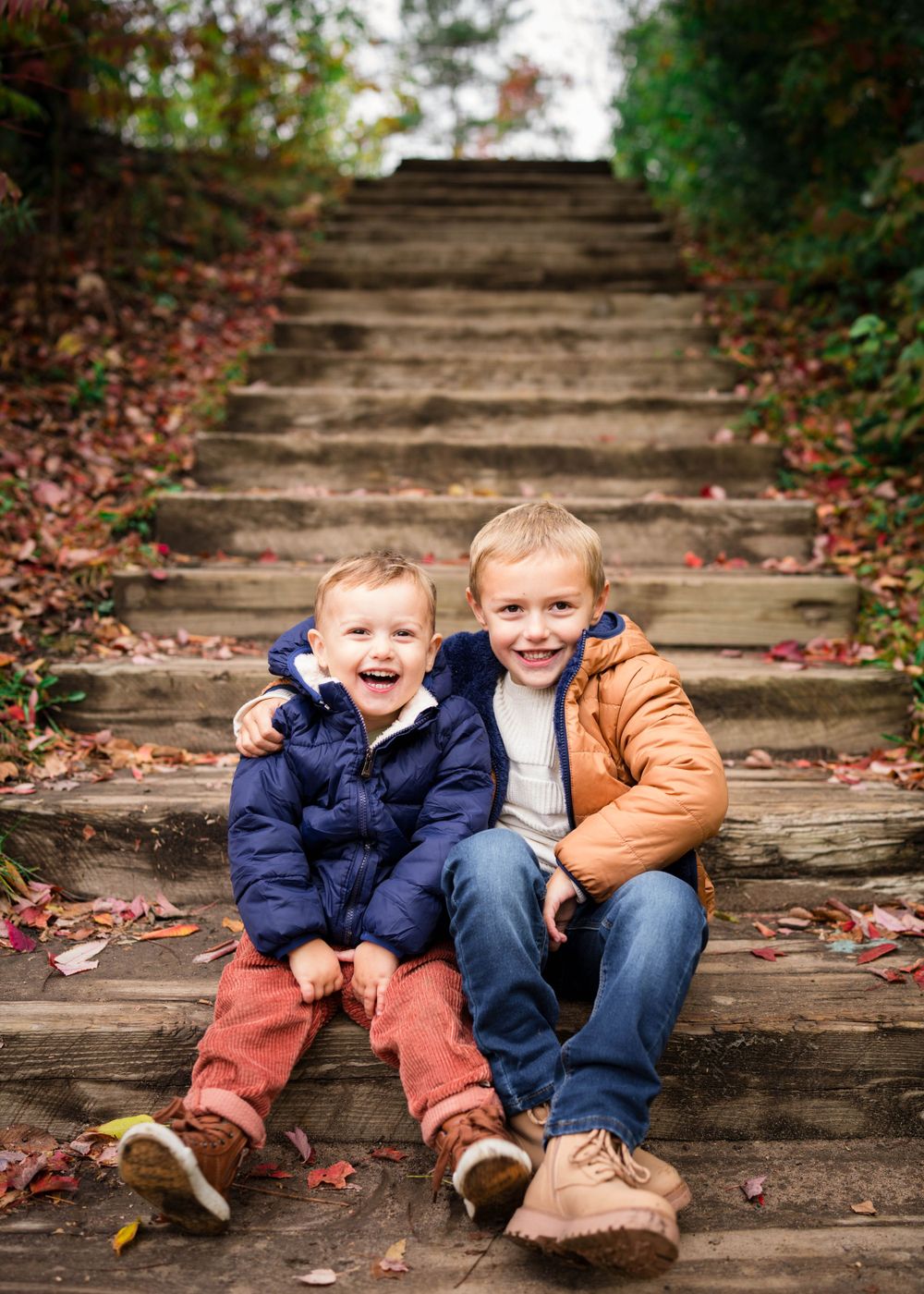 Two young boys in colorful winter jackets laugh and smile sitting on rustic wooden outdoor stairs covered in fall leaves.