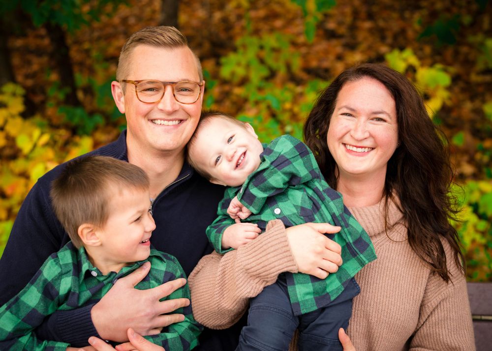 Happy family of four with two young boys in matching green plaid shirts smiling outdoors among golden autumn foliage.