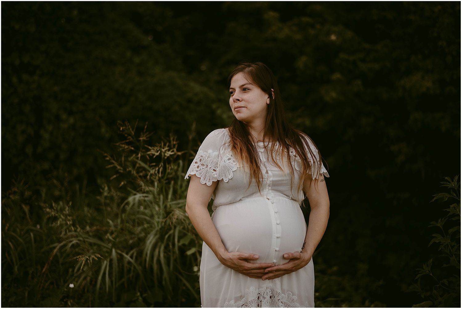 Serene maternity portrait in a white lace dress against lush garden greenery.