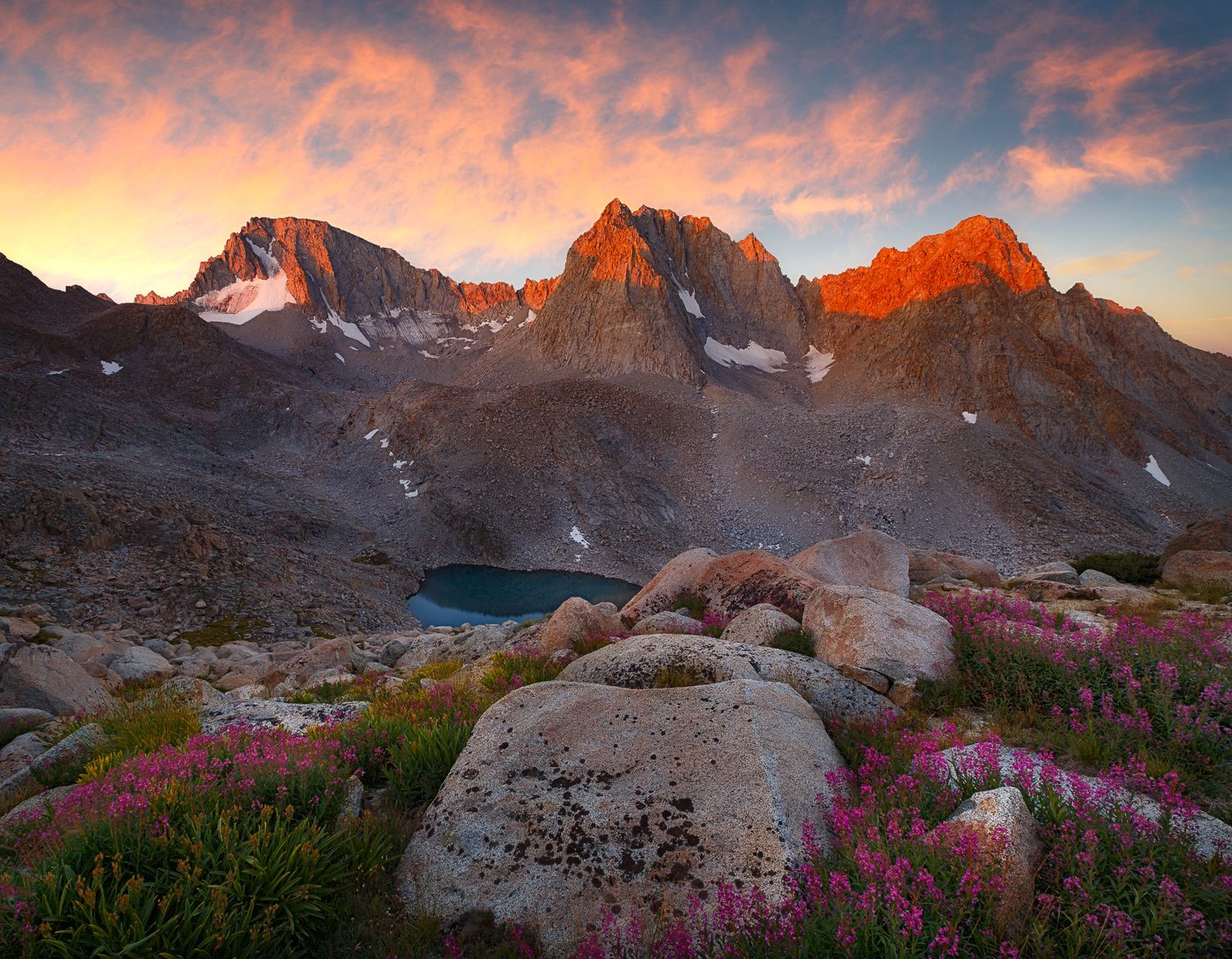 Dramatic mountain peaks glow orange at sunset above an alpine lake with pink wildflowers and granite boulders in the foreground.