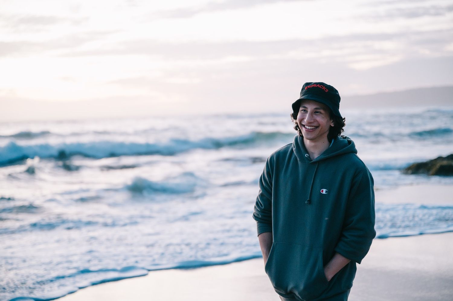 Person in hoodie and beanie smiles while standing on a beach with crashing waves in the background.
