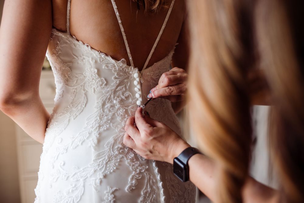 Detailed shot of helping hands buttoning up intricate lace wedding dress.