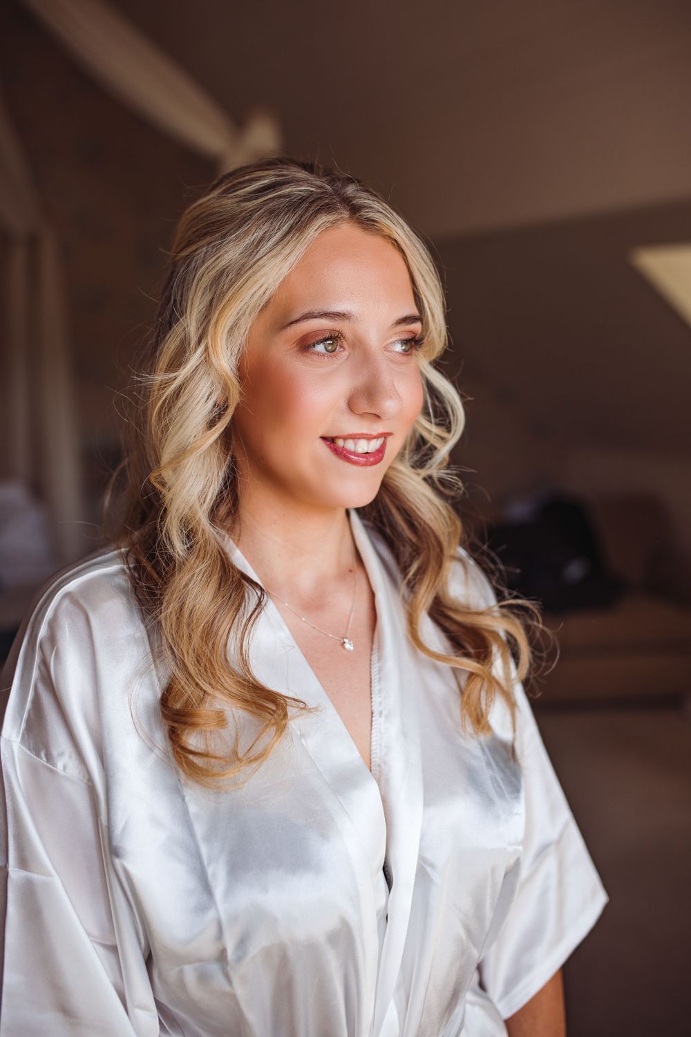 Bride in white silk robe with elegant curled hairstyle and natural makeup getting ready.