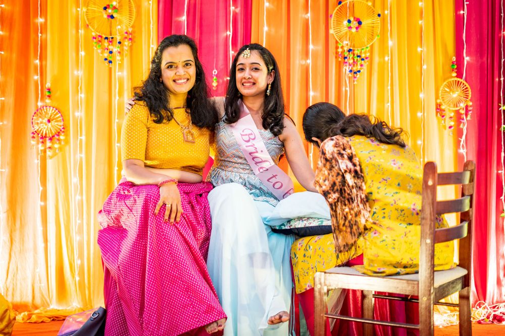 Two women in colorful traditional attire sitting together against vibrant yellow and pink draped backdrop during celebration.