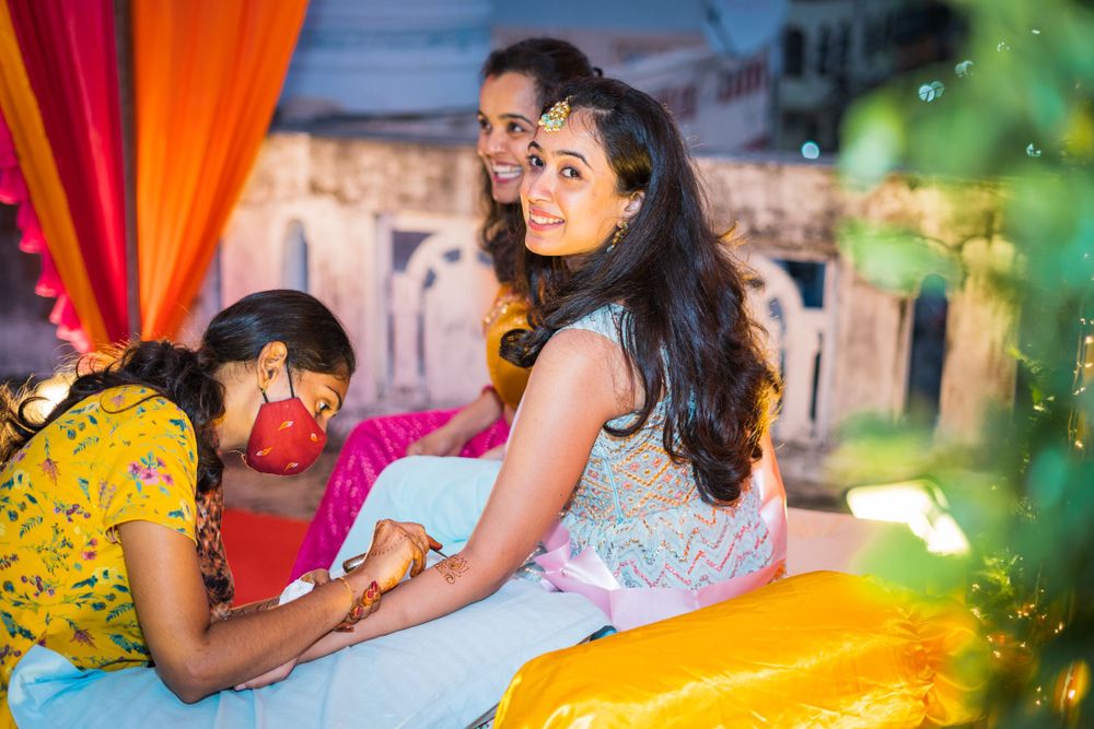 Traditional henna application ceremony with festive orange and red decor in background.