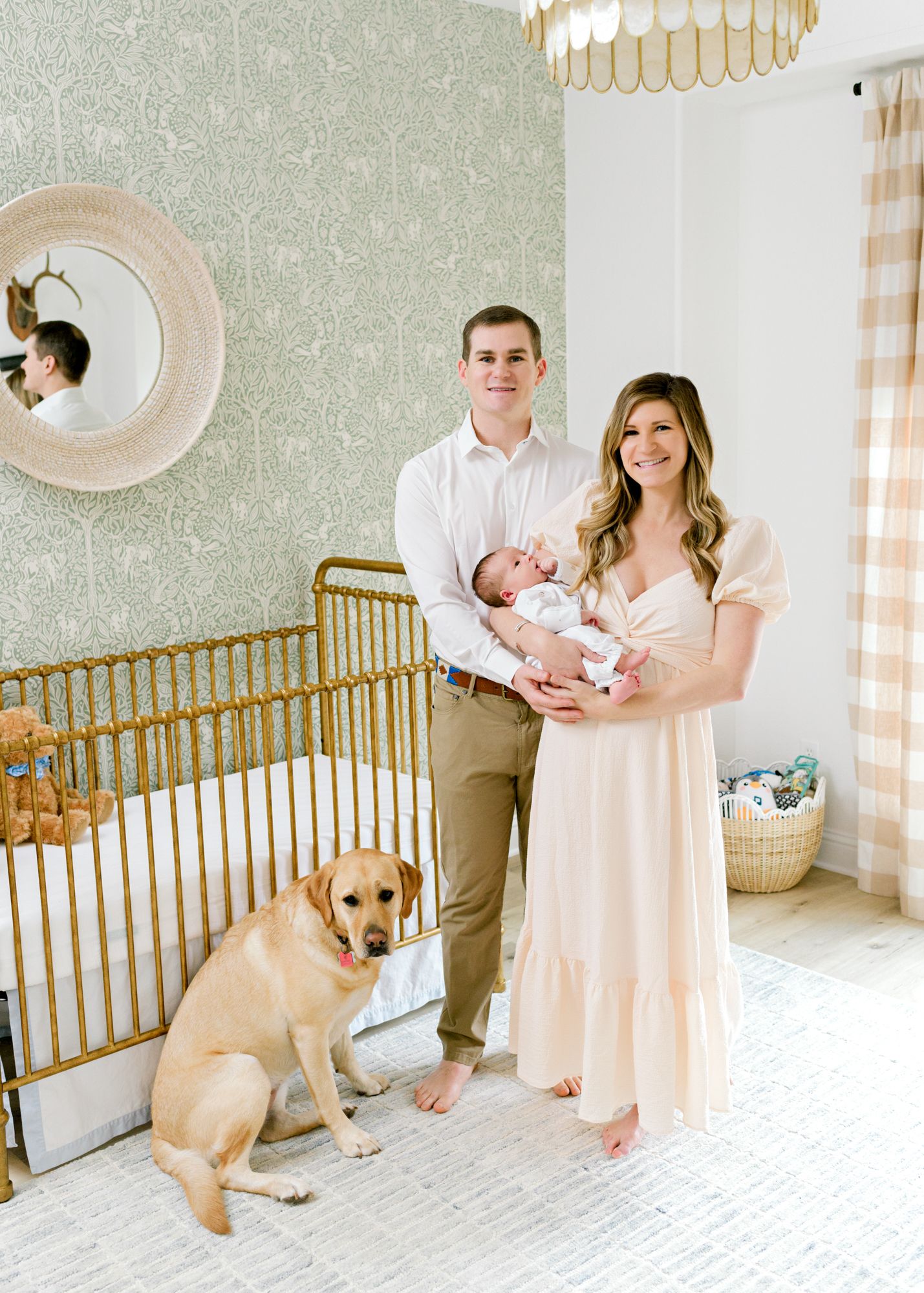 A couple stands in a nursery with their newborn baby and golden retriever beside a gold crib.