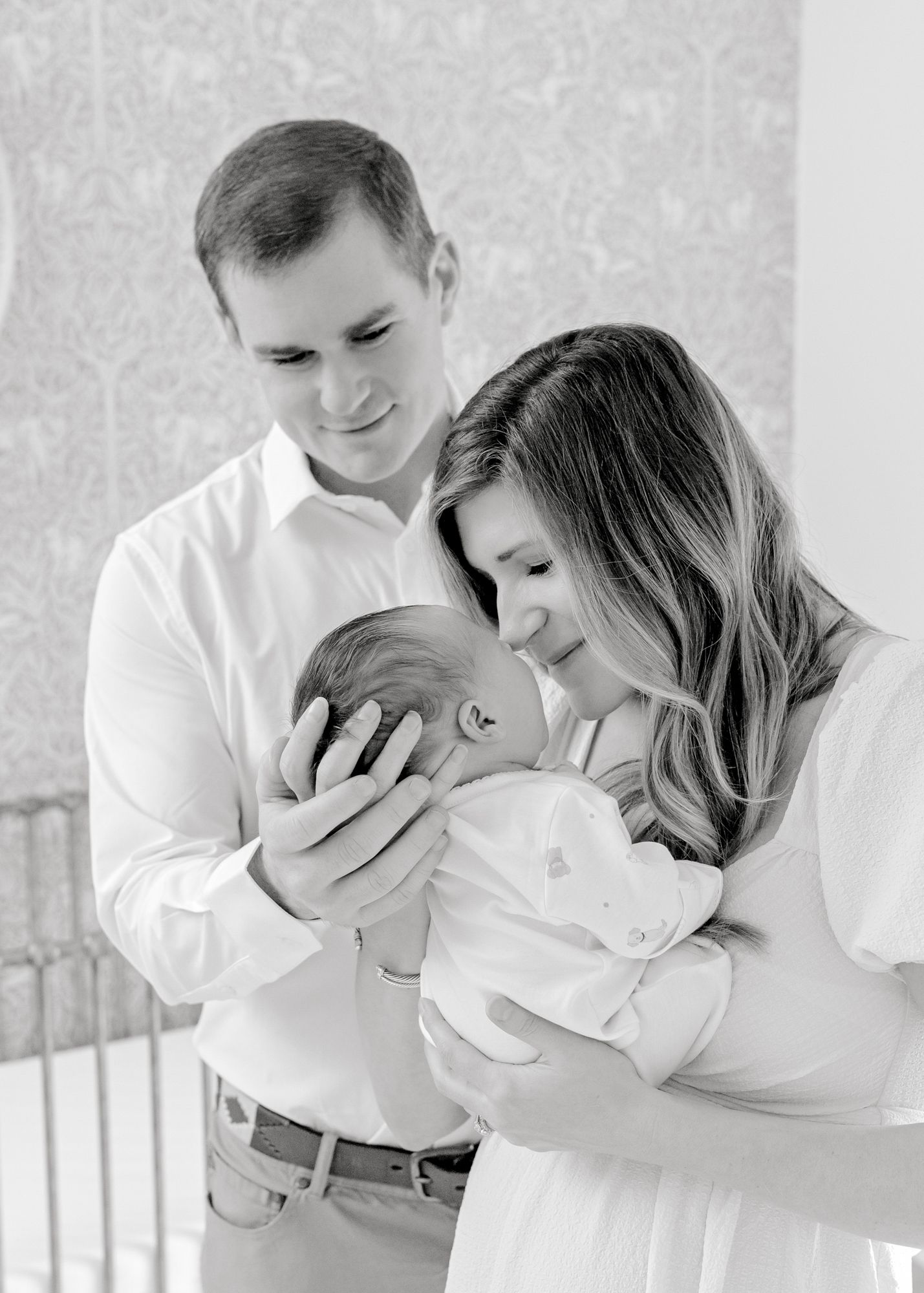 An intimate black and white portrait of parents gazing at their newborn in a nursery setting.