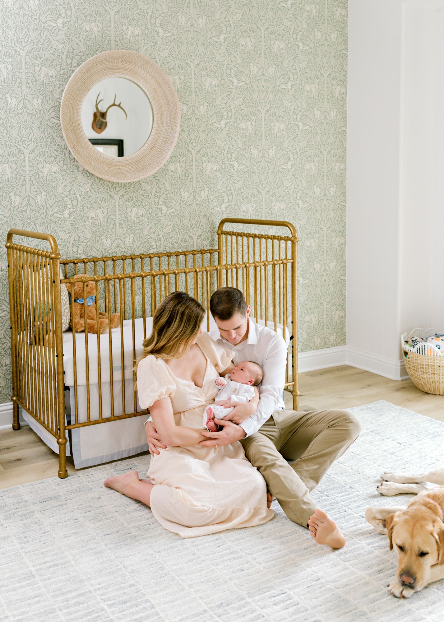 Parents sit by gold crib with newborn baby while golden retriever rests on white carpet.