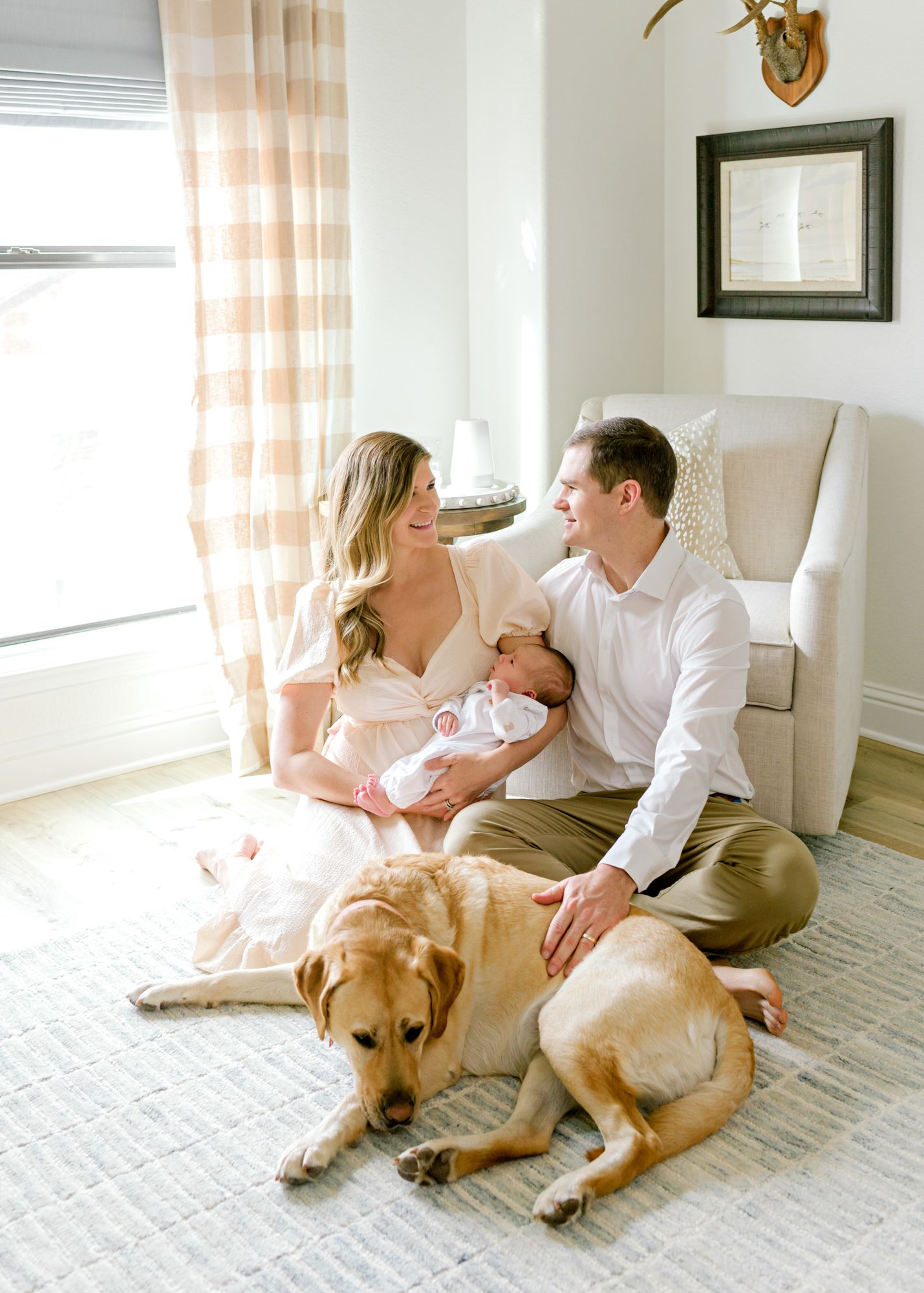 Family sits on cream carpet with newborn baby as golden retriever lounges beside them.