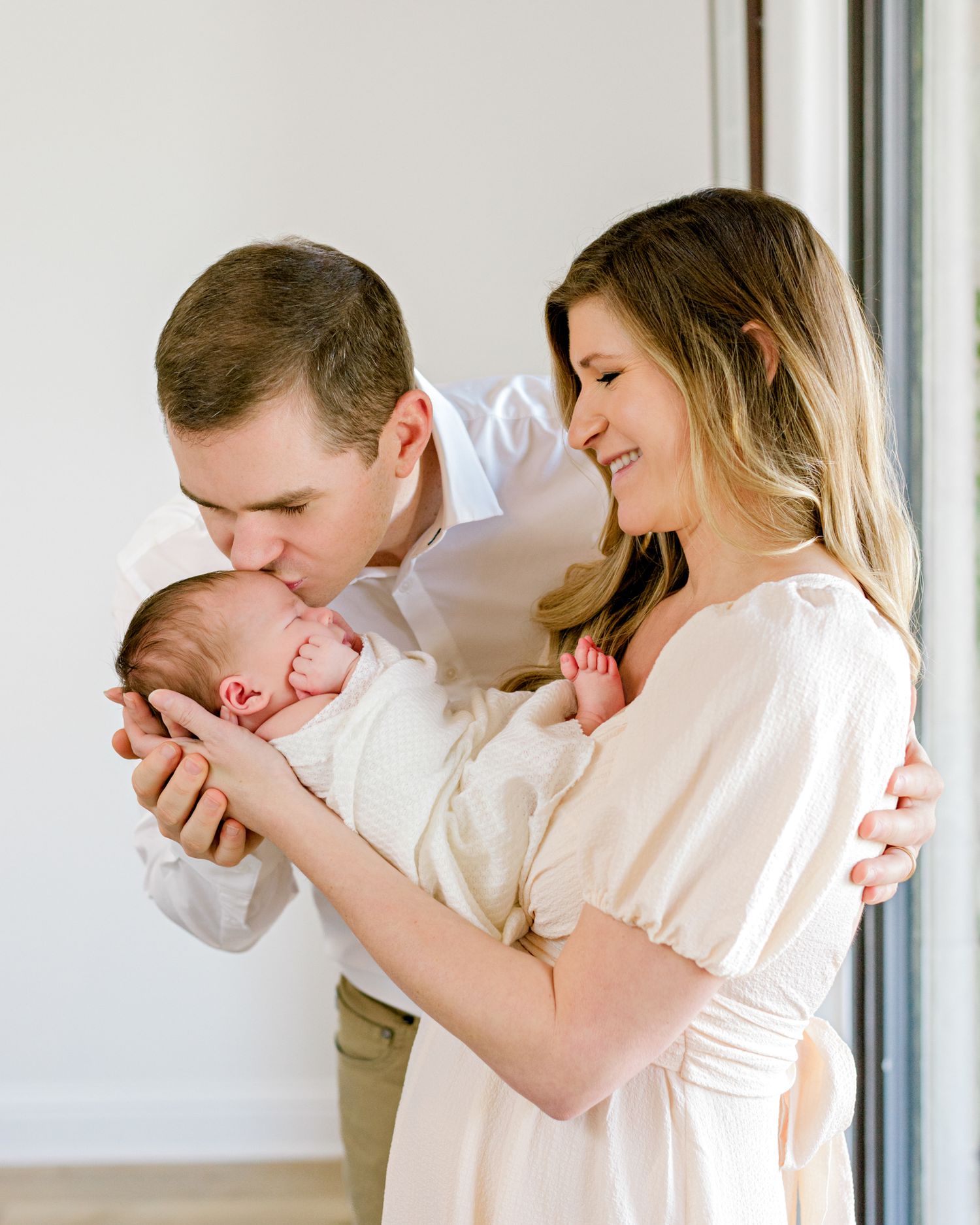 New parents lovingly hold and admire their newborn baby in a tender moment near a window in soft natural light.