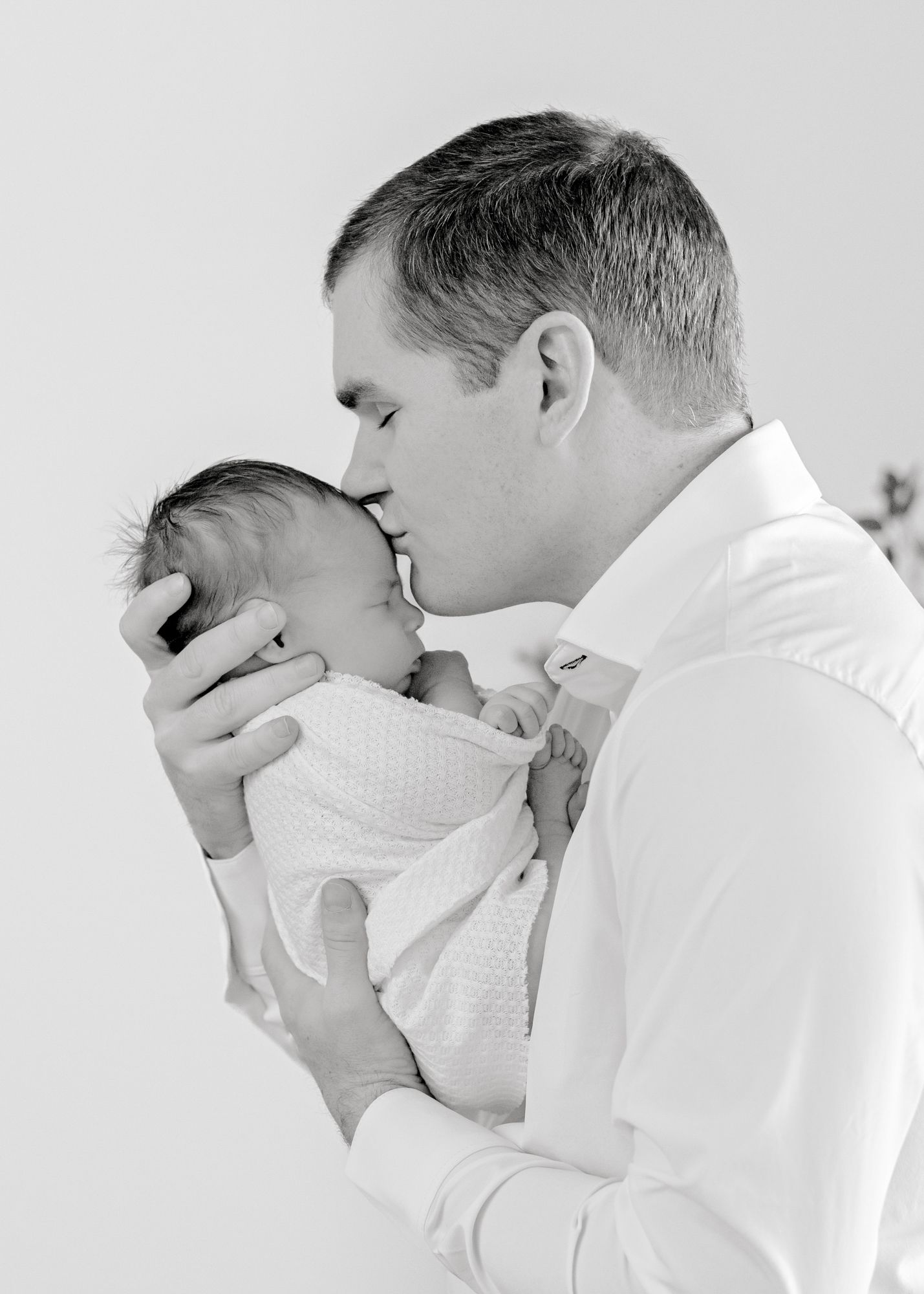Intimate black and white portrait of parent giving newborn baby gentle forehead kiss.