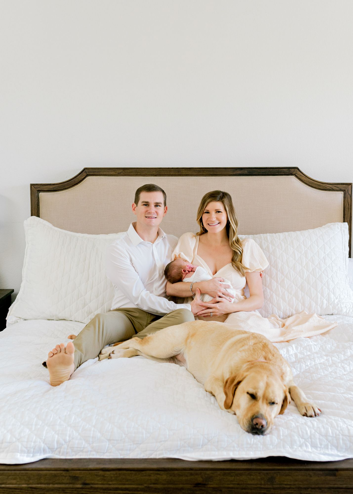 Family relaxes on white bed with golden retriever and newborn baby.