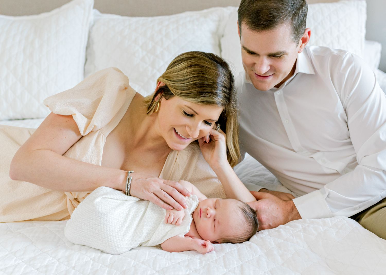 Parents gaze lovingly at newborn baby on white bedding.