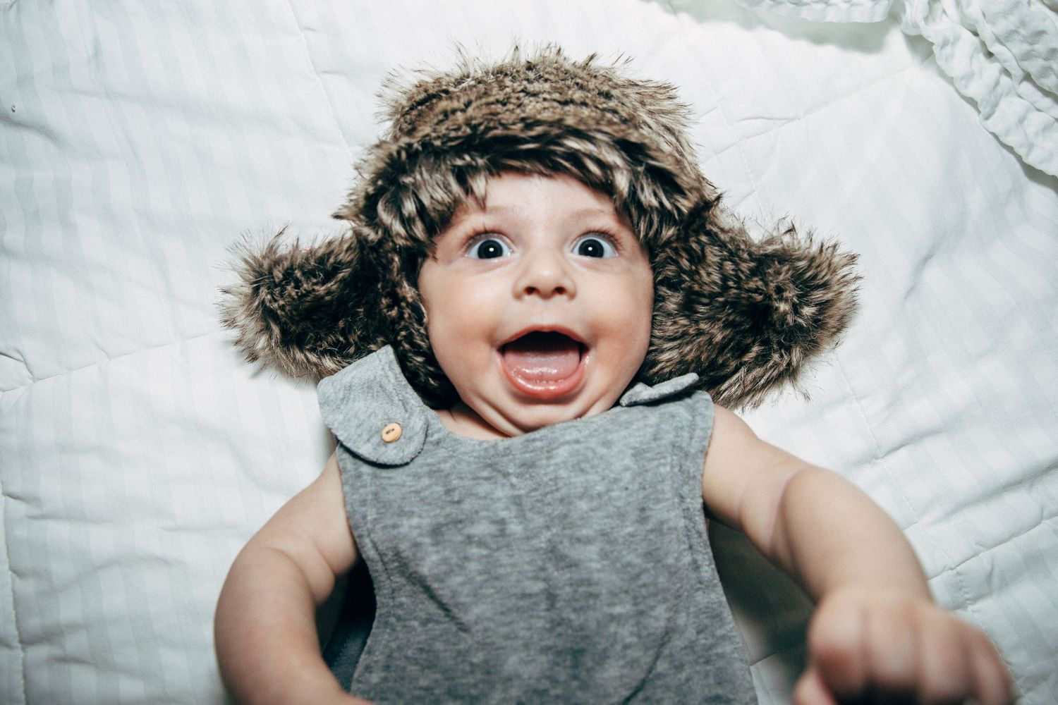 Baby wearing grey shirt and furry winter hat smiles with excited expression.