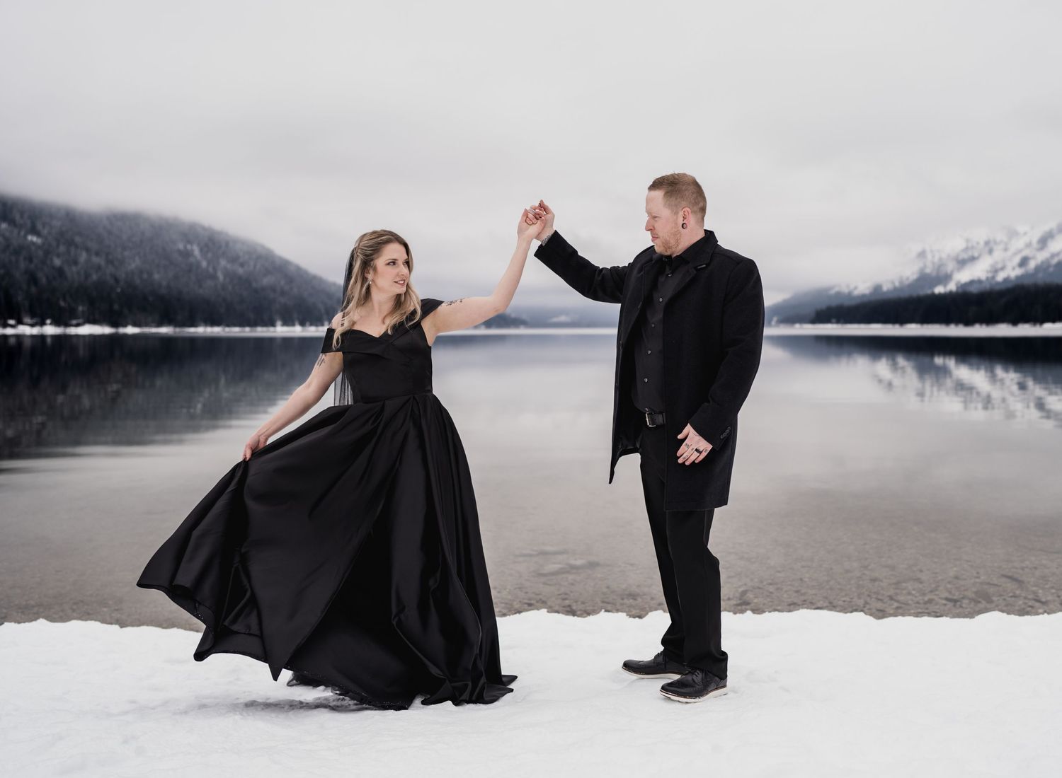 A couple dances in formal black attire on a snowy beach with mountains in the background.