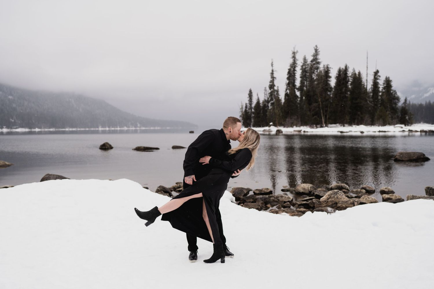 A romantic dip pose on a snowy lakeshore with evergreen trees and mountains in the distance.