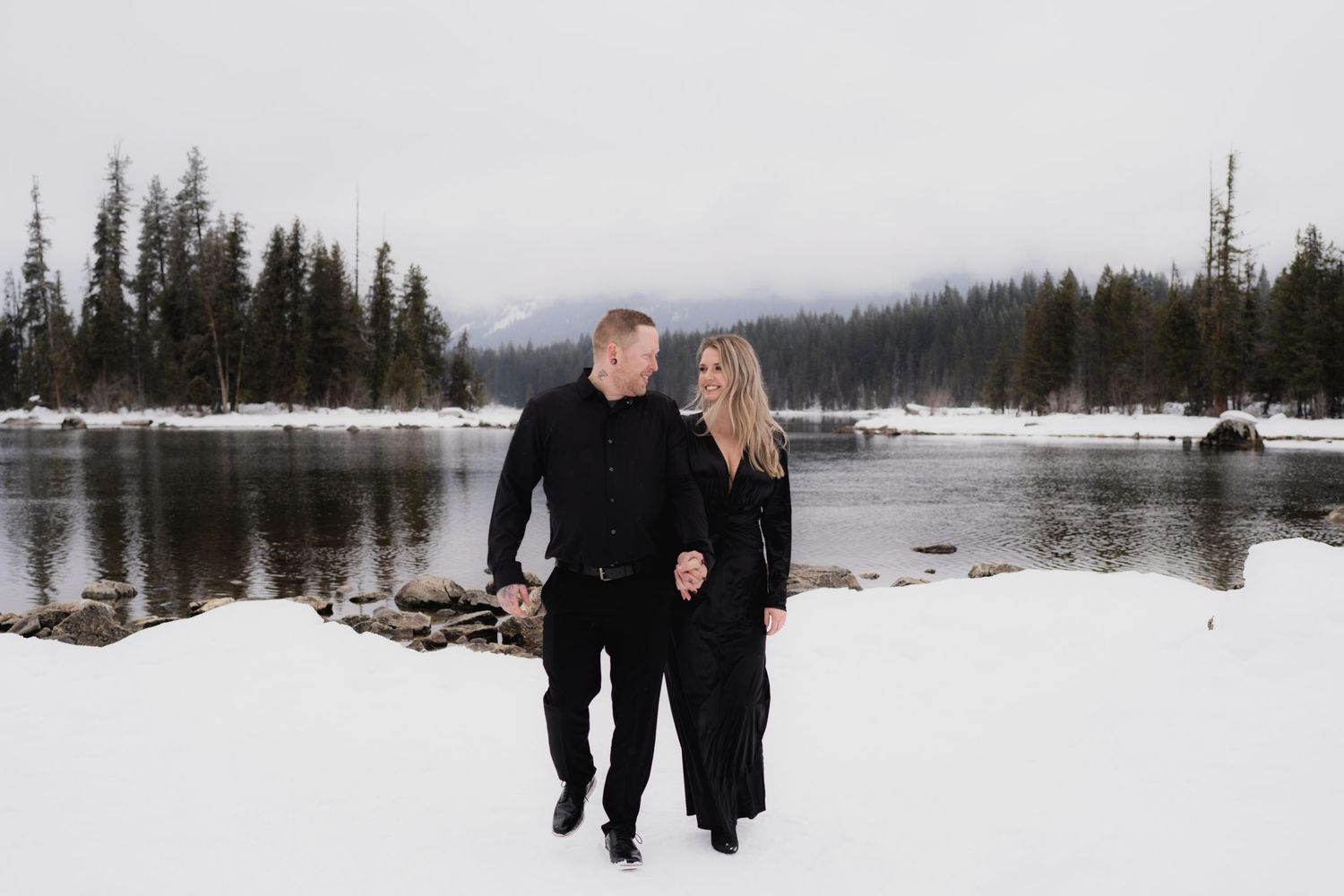 A couple walks together on a snowy shoreline with evergreen trees and mountains in the background.