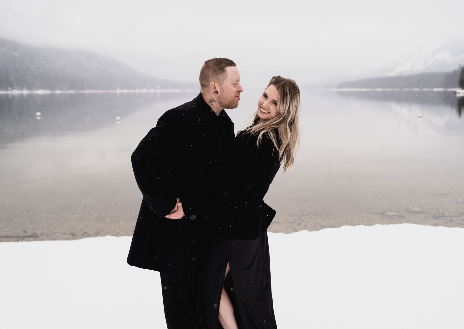 A couple shares an intimate moment by a misty lake surrounded by snow and mountains.