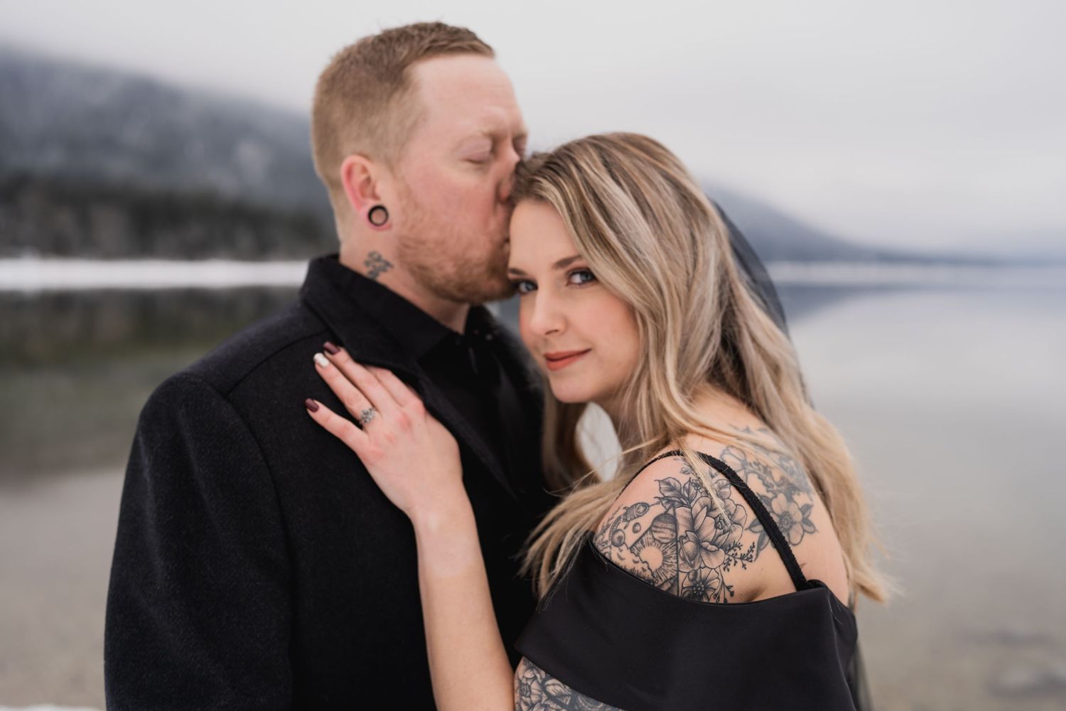 A couple poses intimately against a misty mountain lake backdrop in winter.