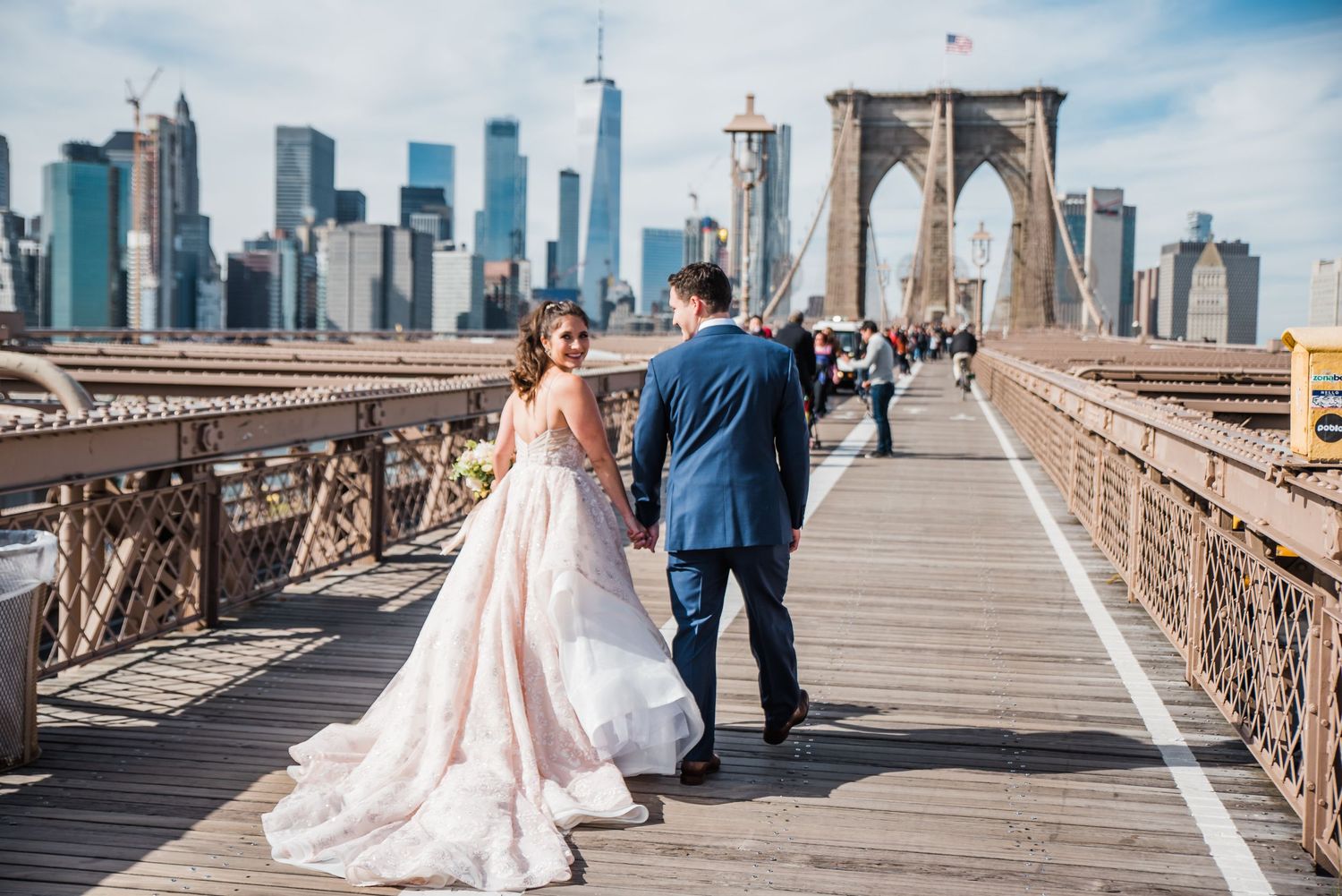 Bride's dress flows in the wind as couple walks on Brooklyn Bridge with Manhattan skyline in background.