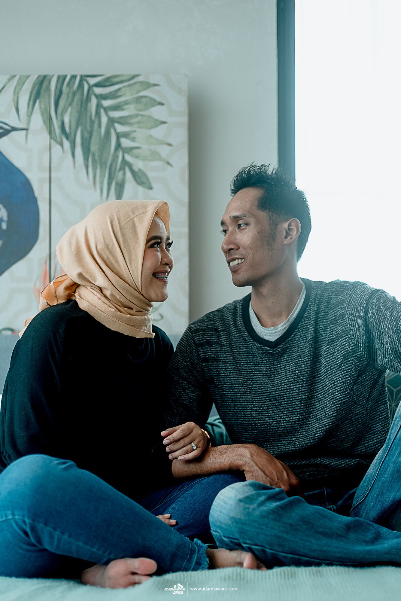 Casual indoor portrait of couple sitting on couch with tropical wall art in background.