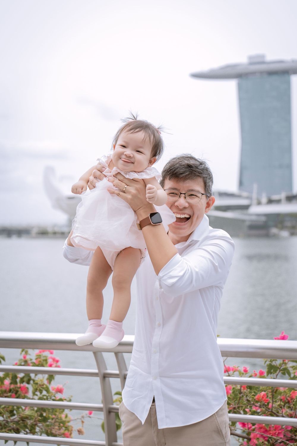 A baby in white dress being lifted up with Marina Bay Sands visible in background.