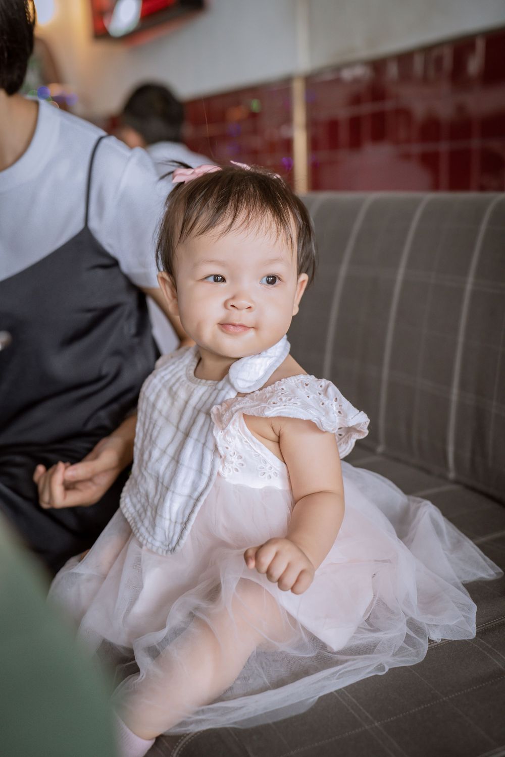 A baby in a white tulle dress smiles while sitting on a grey couch.