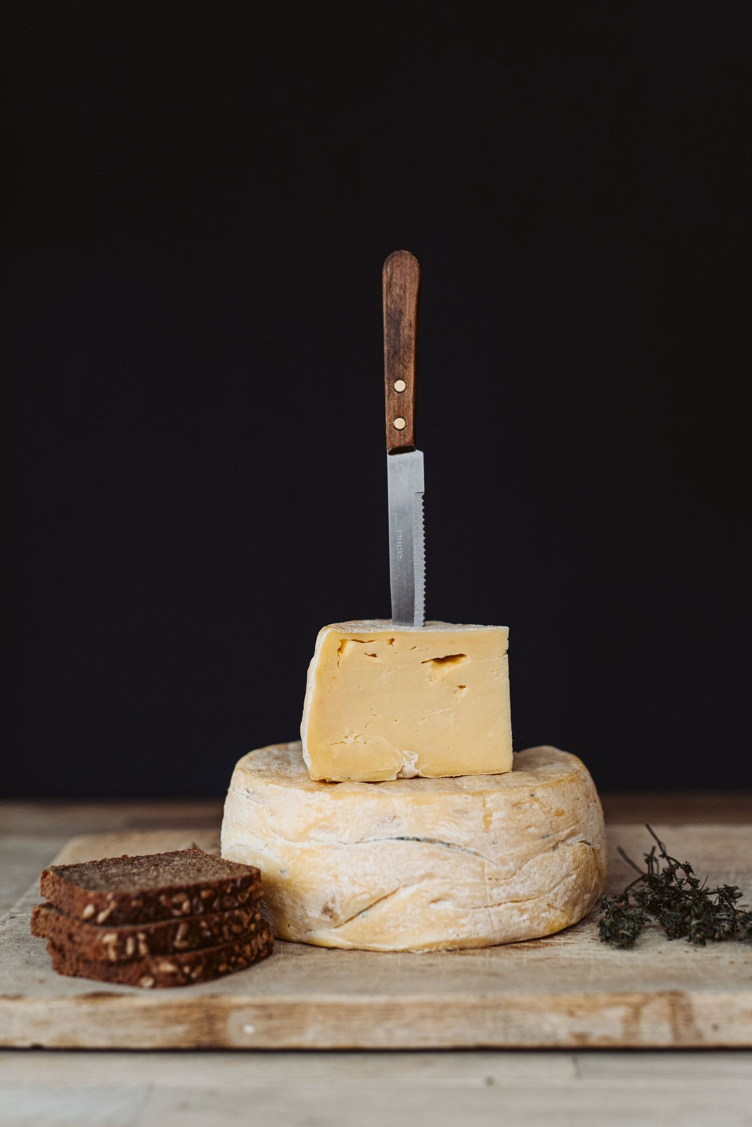 Cheese knife inserted into a stack of aged cheese wheels with dark bread on rustic wooden board.