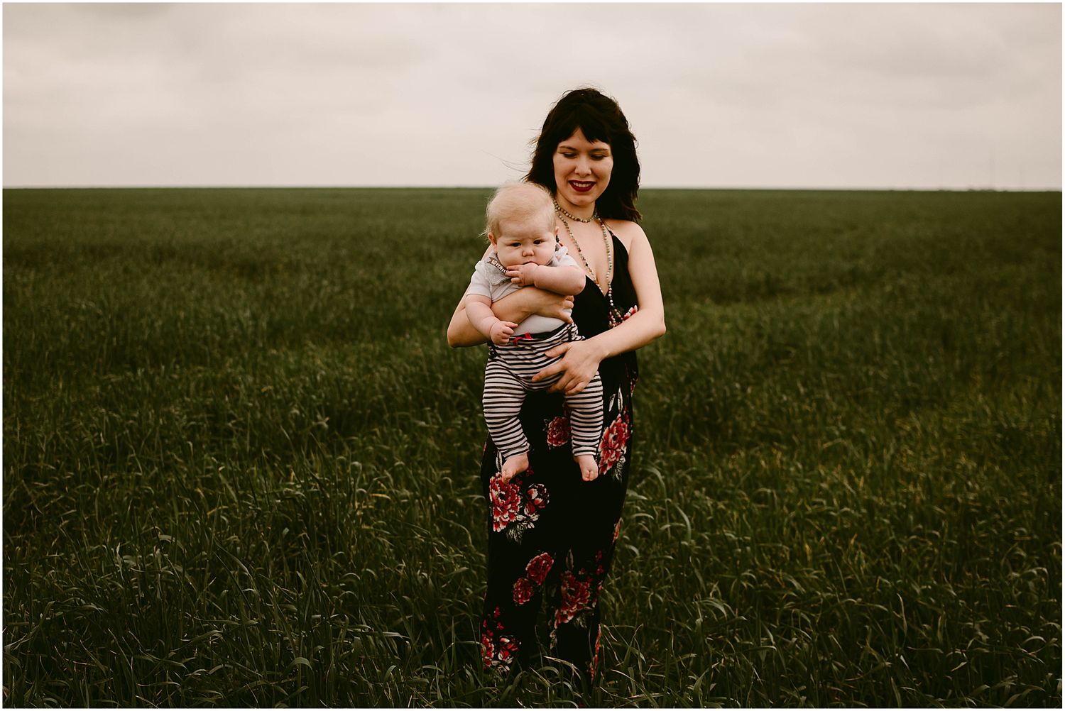 Adult in floral pants holds baby while standing in a serene meadow under cloudy skies.